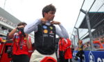 Formula 1 driver in a Ferrari pit vest adjusting his collar before a race, with crew and trackside barriers in the background
