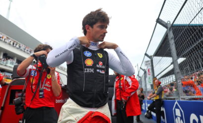 Formula 1 driver in a Ferrari pit vest adjusting his collar before a race, with crew and trackside barriers in the background