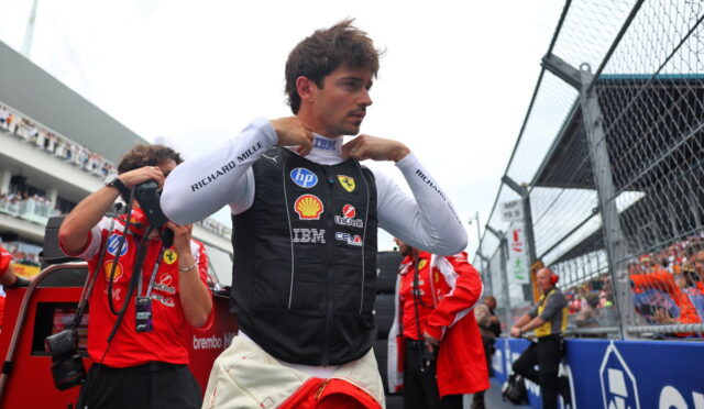 Formula 1 driver in a Ferrari pit vest adjusting his collar before a race, with crew and trackside barriers in the background