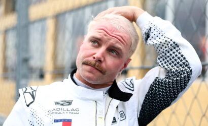 Racer in a white Cadillac Formula 1 team suit touches his head while standing beside a chain-link fence, outdoors