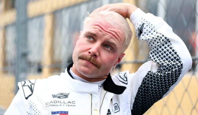 Racer in a white Cadillac Formula 1 team suit touches his head while standing beside a chain-link fence, outdoors