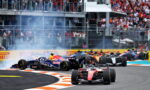 Red Ferrari Formula 1 car leads a pack around a curved track as smoke rises and a crowded grandstand watches.