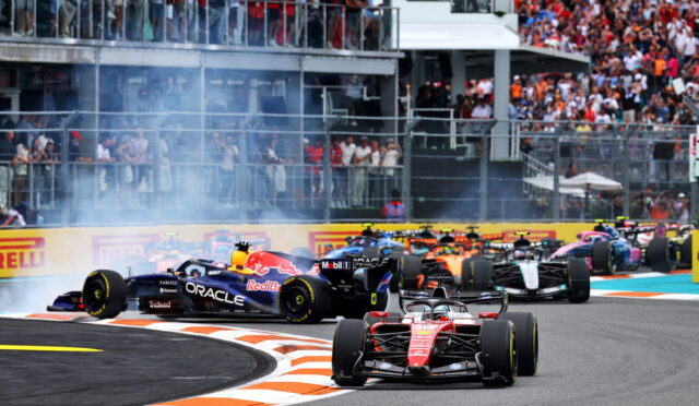 Red Ferrari Formula 1 car leads a pack around a curved track as smoke rises and a crowded grandstand watches.