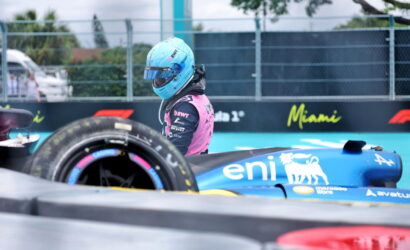 Race car driver in a pink suit and blue helmet beside a blue EnI Formula 1 car on a Miami track yard.