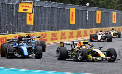 Formula 1 cars race side by side on a track with DHL banners along the barriers.