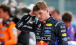 Race car driver in a dark blue sponsor-filled racing suit, adjusting his hair in the pit area.