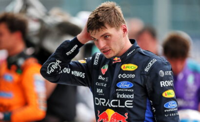 Race car driver in a dark blue sponsor-filled racing suit, adjusting his hair in the pit area.