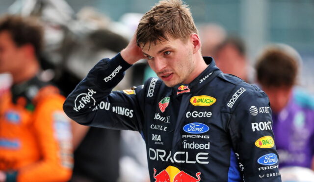 Race car driver in a dark blue sponsor-filled racing suit, adjusting his hair in the pit area.