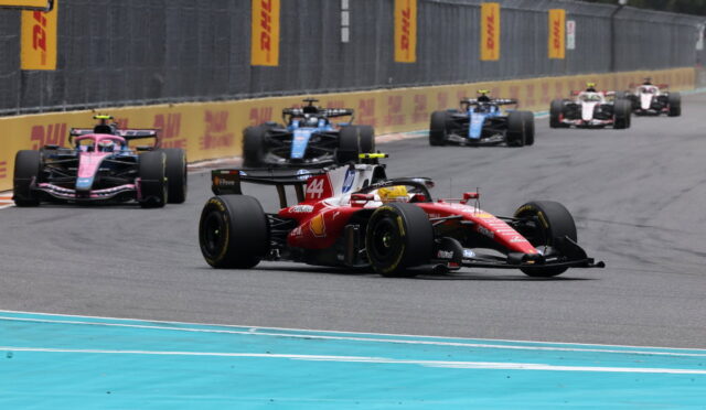 Red Formula 1 car leads a pack on a race track with DHL banners along the barriers.