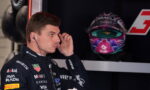 Side profile of a race car driver in a branded suit adjusting an earpiece, with a colorful helmet on display in the background.