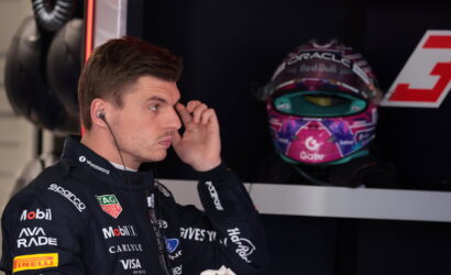 Side profile of a race car driver in a branded suit adjusting an earpiece, with a colorful helmet on display in the background.