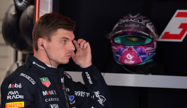 Side profile of a race car driver in a branded suit adjusting an earpiece, with a colorful helmet on display in the background.