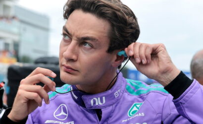 Man in a purple racing suit with sponsor logos, inserting a blue earbud into his ear.