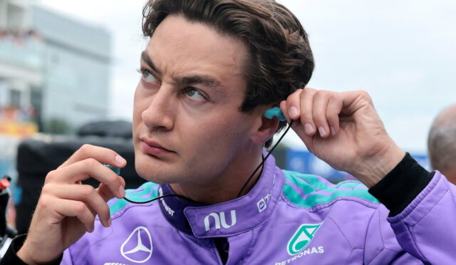 Man in a purple racing suit with sponsor logos, inserting a blue earbud into his ear.