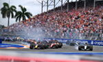Formula 1 cars racing on a circuit with a packed grandstand in the background and fencing along the track.