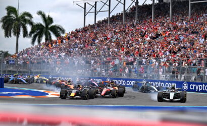 Formula 1 cars racing on a circuit with a packed grandstand in the background and fencing along the track.
