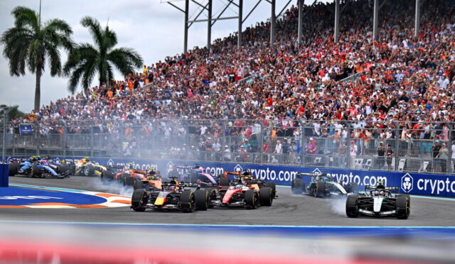 Formula 1 cars racing on a circuit with a packed grandstand in the background and fencing along the track.