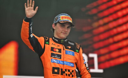 Racing driver in an orange suit raises a hand in victory on a podium, wearing a Pirelli cap and sponsor logos visible on the suit.