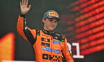 Young race car driver in an orange suit and Pirelli cap waving to the crowd on a podium in celebration.