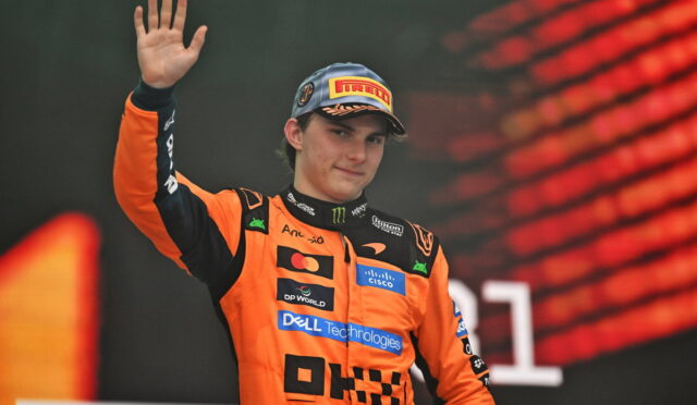 Young race car driver in an orange suit and Pirelli cap waving to the crowd on a podium in celebration.