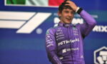 Young Formula One driver in a purple Petronas racing suit on a podium, hand running through hair after a race, smiling at camera.