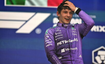 Young Formula One driver in a purple Petronas racing suit on a podium, hand running through hair after a race, smiling at camera.