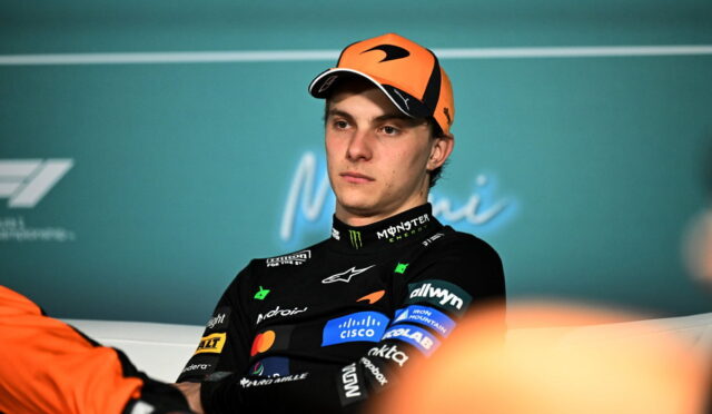 Portrait of a young Formula 1 driver in an orange cap and black racing suit with sponsor logos, seated at a press event.