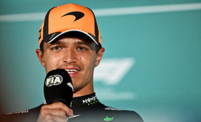 Male race driver in an orange cap speaking into a FIA microphone during a post-race interview.