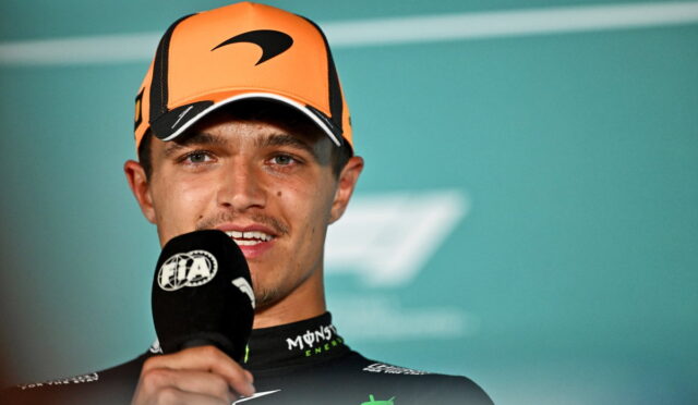 Male race driver in an orange cap speaking into a FIA microphone during a post-race interview.