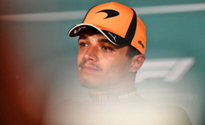 Portrait of a young man wearing an orange cap and dark racing suit, looking to the left with a neutral expression.