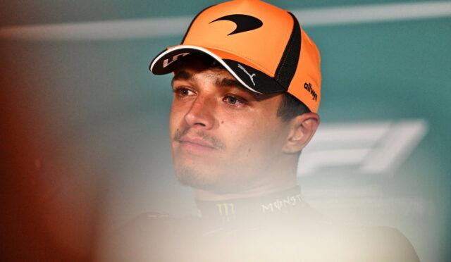 Portrait of a young man wearing an orange cap and dark racing suit, looking to the left with a neutral expression.