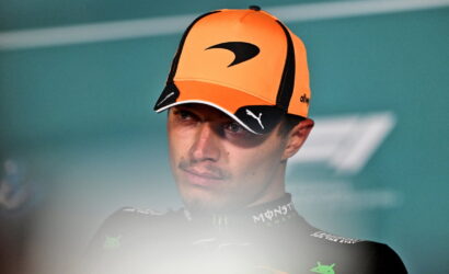 Portrait of a male race driver wearing an orange cap and a dark racing suit, looking to the left.