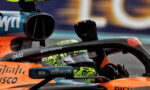 Close-up of an orange Formula 1 car cockpit with a driver raising a gloved fist, neon-green helmet visible in the seat area, sponsor logos shown.