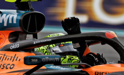 Close-up of an orange Formula 1 car cockpit with a driver raising a gloved fist, neon-green helmet visible in the seat area, sponsor logos shown.