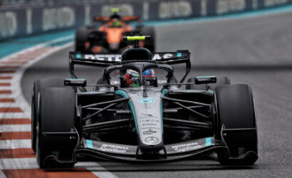 Front view of a Mercedes F1 car leading a race, racing on a track with red-and-white curbs in the foreground, sponsor logos visible on the car body.