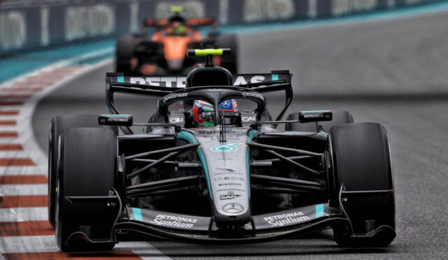 Front view of a Mercedes F1 car leading a race, racing on a track with red-and-white curbs in the foreground, sponsor logos visible on the car body.