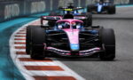 Front view of a pink and blue Formula 1 car racing on a curved track with red-and-white barriers in the background