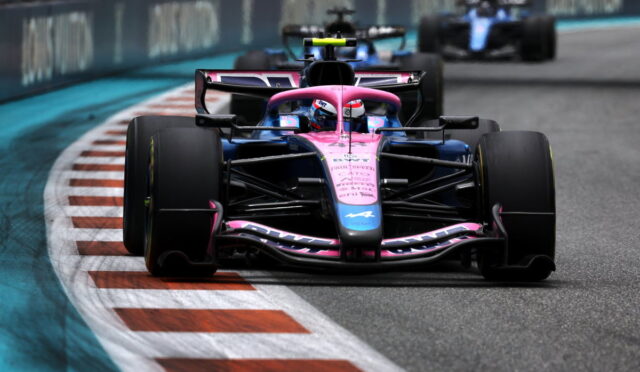 Front view of a pink and blue Formula 1 car racing on a curved track with red-and-white barriers in the background