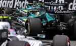Teal Formula 1 race car in a crowded pit area, surrounded by other cars and sponsor logos like Valvoline and Coinbase.