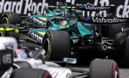Teal Formula 1 race car in a crowded pit area, surrounded by other cars and sponsor logos like Valvoline and Coinbase.