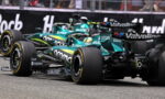 Two teal Formula 1 cars race side by side on a track, with sponsor logos like Coinbase and Valvoline visible.