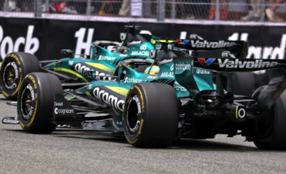Two teal Formula 1 cars race side by side on a track, with sponsor logos like Coinbase and Valvoline visible.