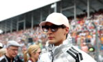 Man wearing a light pink cap, dark sunglasses and a shiny silver racing jacket at a motorsport event.
