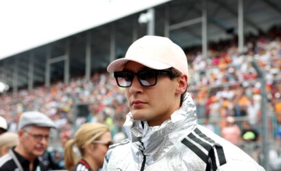 Man wearing a light pink cap, dark sunglasses and a shiny silver racing jacket at a motorsport event.