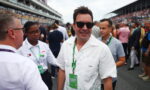 Man in a white patterned shirt and sunglasses smiles at the camera amid a crowded racetrack event.