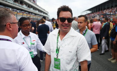 Man in a white patterned shirt and sunglasses smiles at the camera amid a crowded racetrack event.