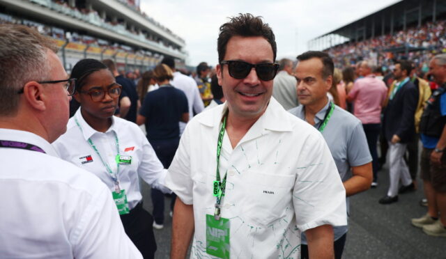 Man in a white patterned shirt and sunglasses smiles at the camera amid a crowded racetrack event.