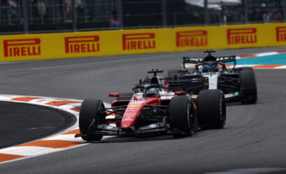 Red Formula 1 car leads a corner, closely followed by another car on a race track with orange-and-white curbs and bright Pirelli boards in the background