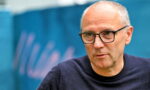 Older man with glasses in a dark sweater, looking to the side, outdoors in front of a blue backdrop.