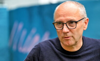 Older man with glasses in a dark sweater, looking to the side, outdoors in front of a blue backdrop.